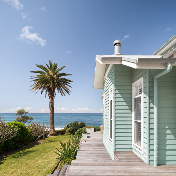 Side elevation of the Wombarra No. 3 home by Simpson & Co Projects, with green weatherboards, white trims, verandah post and ocean views with palm tree.