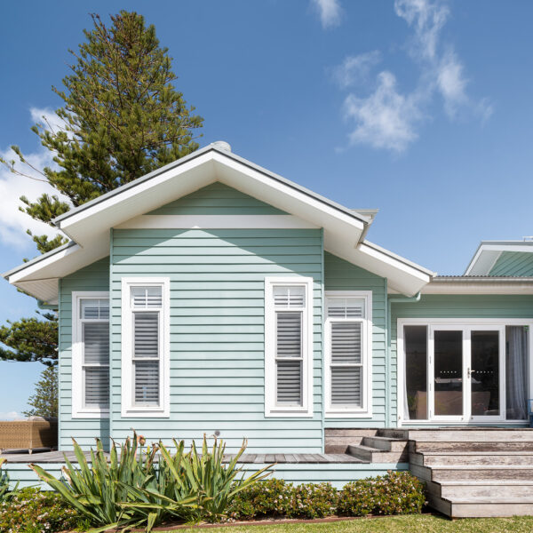 Front elevation of the Wombarra No. 3 home by Simpson & Co Projects, with green weatherboards, white trims, gabled roof and verandah surrounded by coastal trees.
