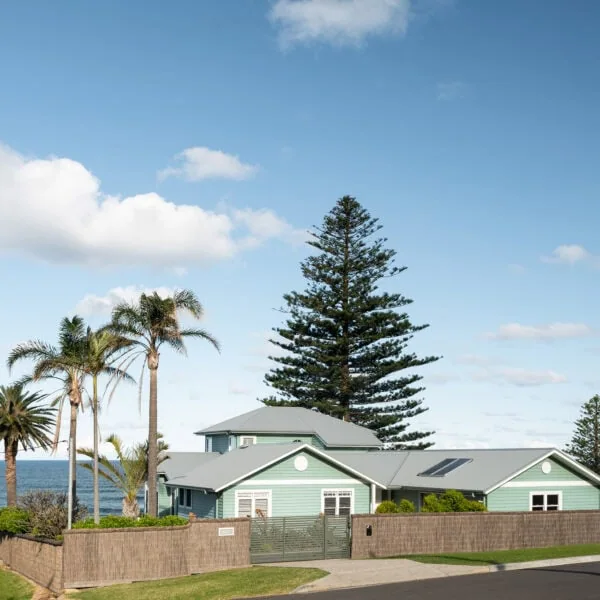 Street-facing coastal elevation of the Wombarra No. 3 home by Simpson & Co Projects, with soft green weatherboards, white trim and a tall Norfolk pine.