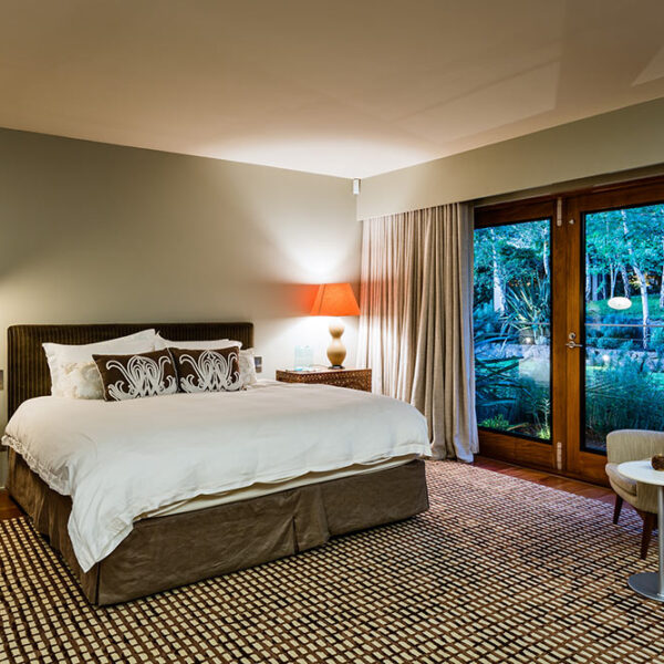 Master bedroom at Ooralba Estate featuring a large bed, soft neutral furnishings, warm lighting, and sliding glass doors overlooking the Kangaroo Valley landscape.