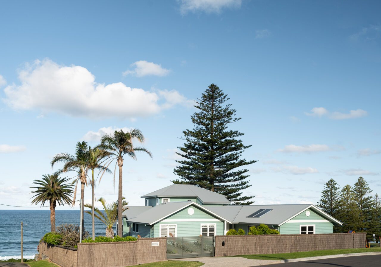 Street-facing coastal elevation of the Wombarra No. 3 home by Simpson & Co Projects, with soft green weatherboards, white trim and a tall Norfolk pine.